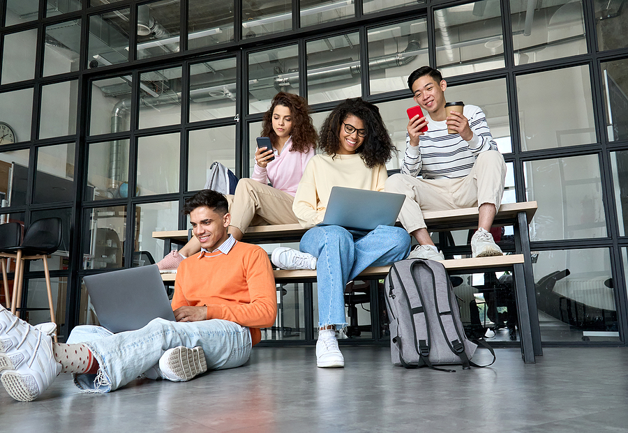 Young happy cheerful multiethnic cool group of high school college students girls and guys sitting at university campus using digital devices social media drinking coffee preparing to seminar classes.